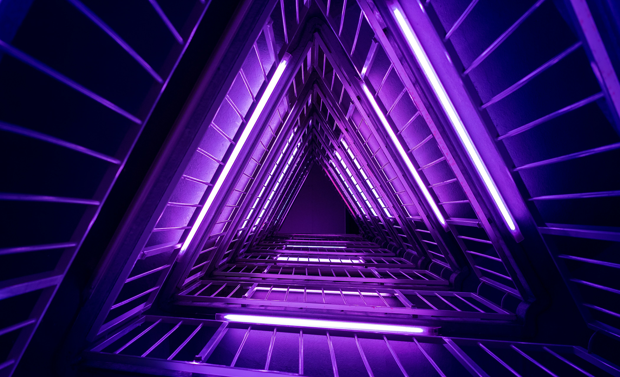 Looking up a stairwell of nested triangular landings lit by purple fluorescent tubes